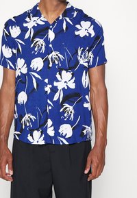 Lyhythihainen paita sinisestä kankasta, jossa on valkoisia kukka- ja mustia lehtikuvioita. Paidassa on napitettava etuosa ja leirikaulus.