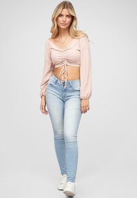 Top a maniche lunghe cropped rosa chiaro con arricciature e dettagli a fiocco, abbinato a jeans skinny blu chiaro e sneakers bianche.