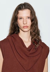 Braunes ärmelloses Top mit drapierter Schalkragen-Design. Weicher Stoff und entspannter Schnitt. Das Model hat welliges Haar und steht vor einem hellen Hintergrund.