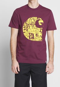 Bordowa bawełniana koszulka z dużym żółtym wzorem graficznym, zawierającym różne logotypy i elementy tekstowe. Klasyczny okrągły dekolt i krótkie rękawy.