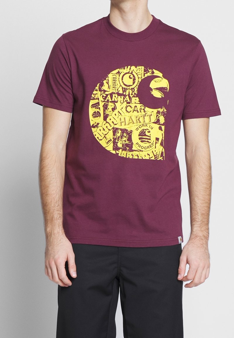 Bordowa bawełniana koszulka z dużym żółtym wzorem graficznym, zawierającym różne logotypy i elementy tekstowe. Klasyczny okrągły dekolt i krótkie rękawy.