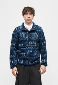 Νεαρός άνδρας που φοράει μπλε φλις πουλόβερ με σχέδιο πάνω από λευκό πουκάμισο με γιακά και μαύρο παντελόνι cargo, στέκεται μπροστά σε απλο φόντο.