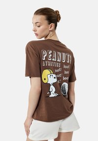 Camiseta de algodón marrón con un estampado gráfico de Snoopy con un casco amarillo, texto que dice "PEANUTS ATHLETICS" y acentos de repetición "¡boot!".