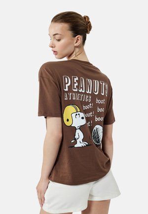 T-shirt en coton marron avec un imprimé graphique de Snoopy portant un casque jaune, avec le texte "PEANUTS ATHLETICS" et des accents répétitifs "boot!".