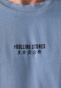 Svijetloplava pamučna majica s okruglim izrezom koja sadrži istaknuti crni tekstualni logo: "THE ROLLING STONES" i dodatne kineske znakove.