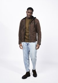 Brun mockaskinnjacka med svart huva, med dragkedjefickor; texturerad grön tröja under; ljusblå jeans; och svarta chelsea-stövlar.