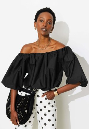Femme portant un top noir épaules dénudées et un pantalon blanc à pois noirs, tenant un sac noir clouté, une main dans la poche.