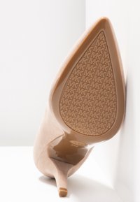 Chaussure à talon haut en daim beige inclinée vers le haut, montrant une semelle à motif avec le logo « MK » répété et une pointe effilée.
