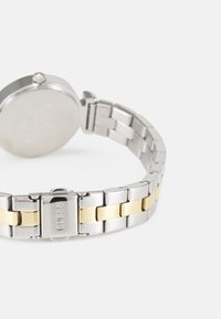 Montre en acier inoxydable avec un boîtier rond, dotée d'un bracelet à maillons argentés et d'accents dorés. Le fermoir est gravé avec "GUESS."