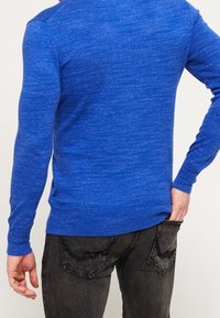 Blauer Langarmpullover mit meliertem Muster, körperbetontem Schnitt und rundem Ausschnitt, kombiniert mit dunklen Jeans mit dekorativen Nähten.