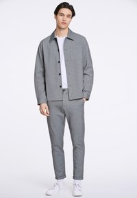 Graue, strukturierte Button-Up-Jacke mit einer Tasche, kombiniert mit passenden maßgeschneiderten Hosen und weißen Sneakern. Schlichtes Design, keine sichtbaren Muster.