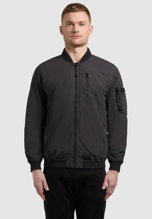 Uomo in piedi, rivolto in avanti, indossa una giacca bomber nera con tasche con cerniera sul petto e sulle maniche, abbinata a pantaloni neri.