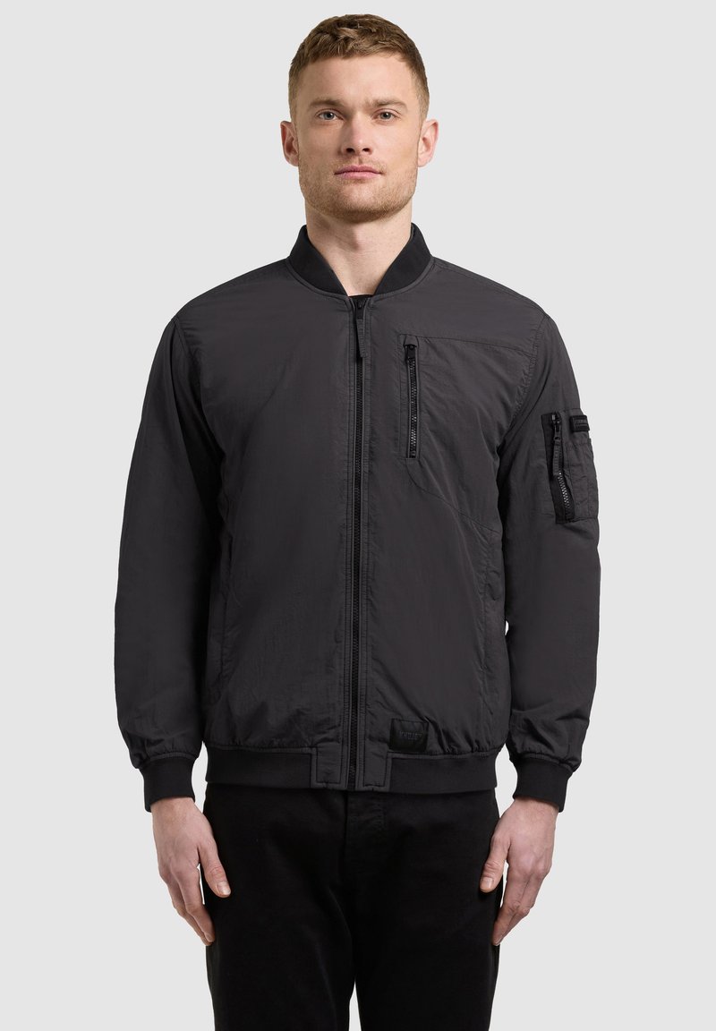 Un homme se tient face à la caméra, portant une veste bomber noire avec des poches zippées sur la poitrine et les manches, associée à un pantalon noir.
