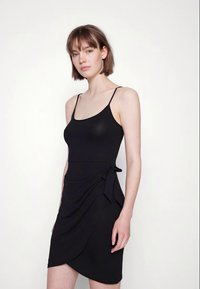 Mulher jovem com cabelo castanho curto usa um vestido preto de alças finas e sem mangas, de pé contra um fundo branco simples.