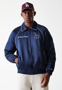 Navy satin jacket with contrasting white piping and embroidered "LA" logo. Features a zip front and elastic cuffs. Brown cap with "LA" logo.