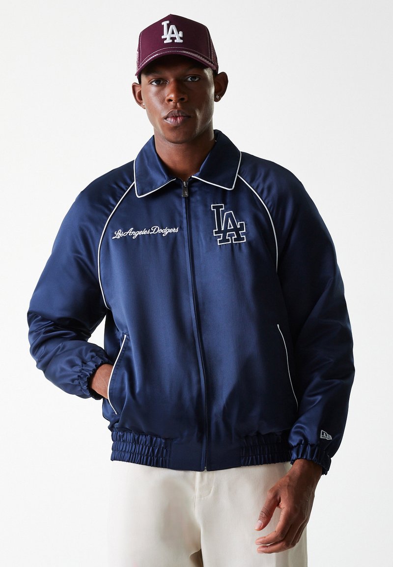 Navy satin jacket with contrasting white piping and embroidered "LA" logo. Features a zip front and elastic cuffs. Brown cap with "LA" logo.