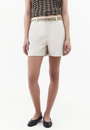 Person mit ärmellosem gemusterten Oberteil, beigefarbenen Shorts mit Gürtel und schwarzen Ballerinas steht vor einfachem weißem Hintergrund.