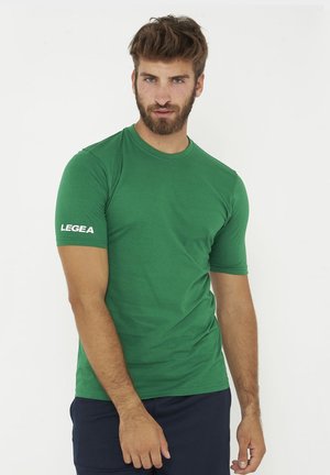 Mann mit kurzem braunem Haar und Bart, der ein grünes LEGEA-T-Shirt und dunkle Hosen vor weißem Hintergrund trägt.