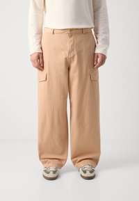 Beige cargo pants with wide legs, featuring two side pockets, a button closure, and a soft, textured fabric. Paired with multicolored sneakers.