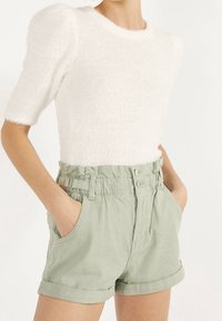 Suéter blanco texturizado con mangas cortas abullonadas, combinado con pantalones cortos de cintura alta en verde claro que presentan dobladillos enrollados y bolsillos laterales.