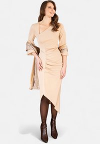 Abito beige con scollo a V, dettaglio arricciato sul lato e orlo inclinato. Accentuato da una sciarpa metallica e abbinato a stivaletti alti marroni.