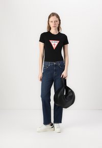 Camiseta negra ajustada con un logotipo triangular rojo "GUESS?", combinada con jeans de pierna recta azul, zapatillas blancas y un bolso negro.