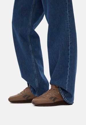 Zapatillas deportivas marrones con detalles en negro, usadas con vaqueros de mezclilla azul sueltos. El diseño del zapato presenta una puntera redonda y una parte superior texturizada.