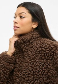 Smešasta smeđa fleece jakna s visokim ovratnikom i zatvaranjem na patentni zatvarač. Tkanina ima luksuznu, hrapavu površinu i duge rukave.