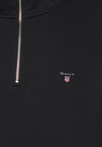 Zwarte zip-up sweatshirt met een zilveren ritssluiting, GANT-logo in het wit en rood op de borst, gemaakt van zachte, gestructureerde stof.