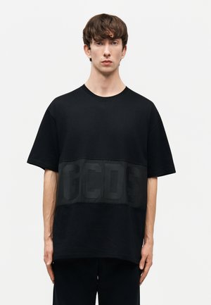 Young man with short brown hair wearing a loose black T-shirt with large embossed "GCDS" letters across the chest, standing against plain background.