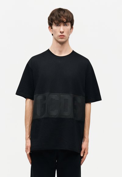Ung man med kort brunt hår som bär en lös svart T-shirt med stora upphöjda bokstäver "GCDS" över bröstet, stående mot en enkel bakgrund.