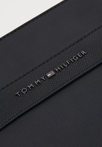 Crna kožna novčanica s teksturiranim završetkom, ugraviranim logom "TOMMY HILFIGER" i prepoznatljivim trobojnim emblemom iznad imena brenda.