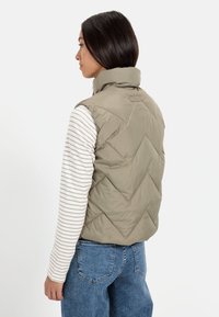 Kaki quilted vest med høj krave, der har et geometrisk mønster, båret over en grå og hvid stribet langærmet skjorte, parret med blå jeans.