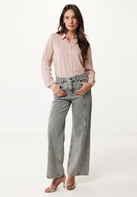 Lichtroze blouse met knopen en lange mouwen, gecombineerd met hoog-genaaide, wijdvallende grijze spijkerbroek. Gouden open-teenschoenen maken de outfit compleet.