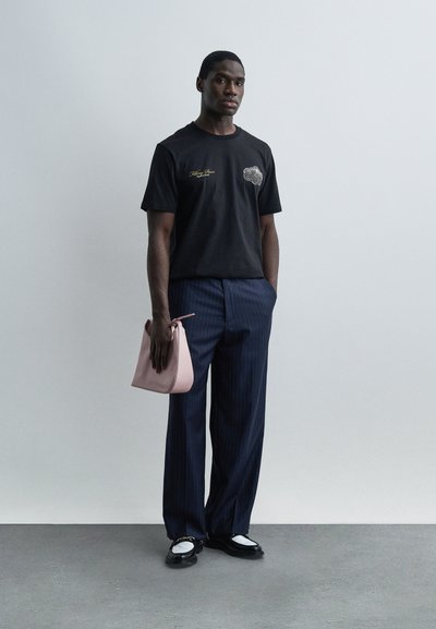 Άνδρας στέκεται μπροστά σε απλό τοίχο φορώντας μαύρο μπλουζάκι με γραφικά, μπλε ναυτικό παντελόνι με λεπτές ρίγες, μαύρα και λευκά μοκασίνια, κρατώντας ροζ παστέλ τσάντα στο πλάι.