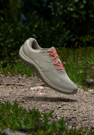 Chaussure de course grise et beige avec des lacets roses flottant au-dessus d’un chemin de gravier entouré de végétation verte.