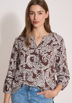 Vrouw draagt een bruin-wit paisley patroon blouse met driekwart mouwen en blauwe jeans, staand met één hand in de zak.