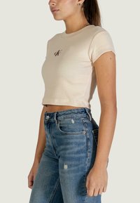 T-shirt beige court en tissu côtelé avec manches courtes et petit logo sur la poitrine, assorti à un jean bleu déchiré taille haute.