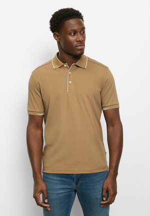 Junger Mann mit kurzen lockigen Haaren, der ein hellbraunes Polohemd mit weißen Akzenten und blaue Jeans trägt, und nach rechts blickt vor einem weißen Hintergrund.