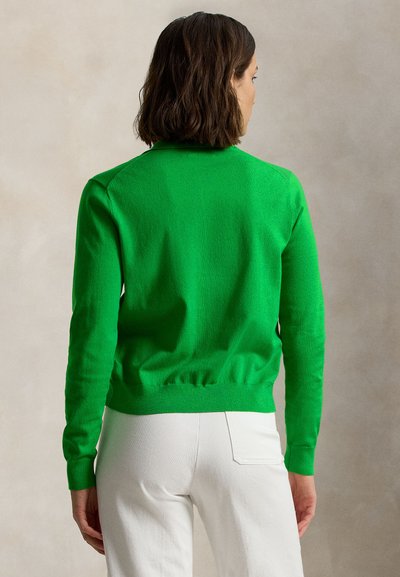 Πράσινο πλεκτό πουλόβερ που φαίνεται από πίσω, με στρογγυλή λαιμόκοψη, ριμπ μανσέτες και εφαρμοστή μέση, συνδυασμένο με λευκό παντελόνι.