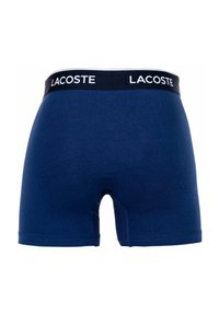 Sötétkék pamut boxeralsó fekete derékkal, amelyen fehér "LACOSTE" felirat található. Az anyag sima, rugalmas textúrájú.