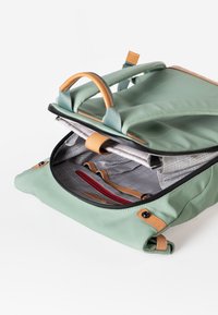 Groene canvas rugzak met tan leren accenten, bovenhandvatten en een ritsopening. Het interieur heeft een geblokt voering en een rode ritszak.