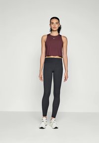 Woman standing wearing a maroon cropped athletic tank, high-waisted black leggings, and white running shoes against a plain light background.