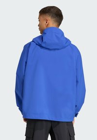 Modrá nepremokavá bunda s kapucňou, voľný strih a dlhé rukávy. Hladká textúra, minimálne detaily, navrhnuté na vonkajšie použitie.