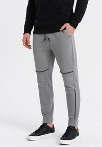 Graue Jogginghose mit schwarzen Akzenten, ausgestattet mit einem taillierten Design, Reißverschlusstaschen und geripptem Material an den Knien. Getragen mit schwarzen Sneakers.