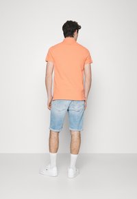 Oranžové polo tričko spárované se světle modrými džíny šortkami, které mají ohrnutý lem. Bílé tenisky a ponožky dotvářejí celý outfit. Pohled zezadu.