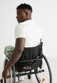 Gyors tolószék fekete szövet ülőrésszel és hát háttámlával, szürke kerekekkel és tároló zsebekkel; a felhasználón világos színű ing és mintás nadrág van.