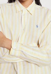 Yellow and white striped button-up shirt made of lightweight fabric, featuring a classic collar and small blue embroidered logo on the chest.