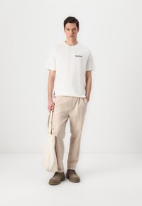 Witte korte mouw t-shirt met blauw logo, beige geplooide broeken, grijze schoenen en een lichtbeige tote bag. Minimalistisch ontwerp en neutrale kleuren.