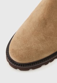 Lysbrune ruskindsstøvler med rund tå, struktureret overflade og en kontrasterende mørkebrun gummisål med et mønster.